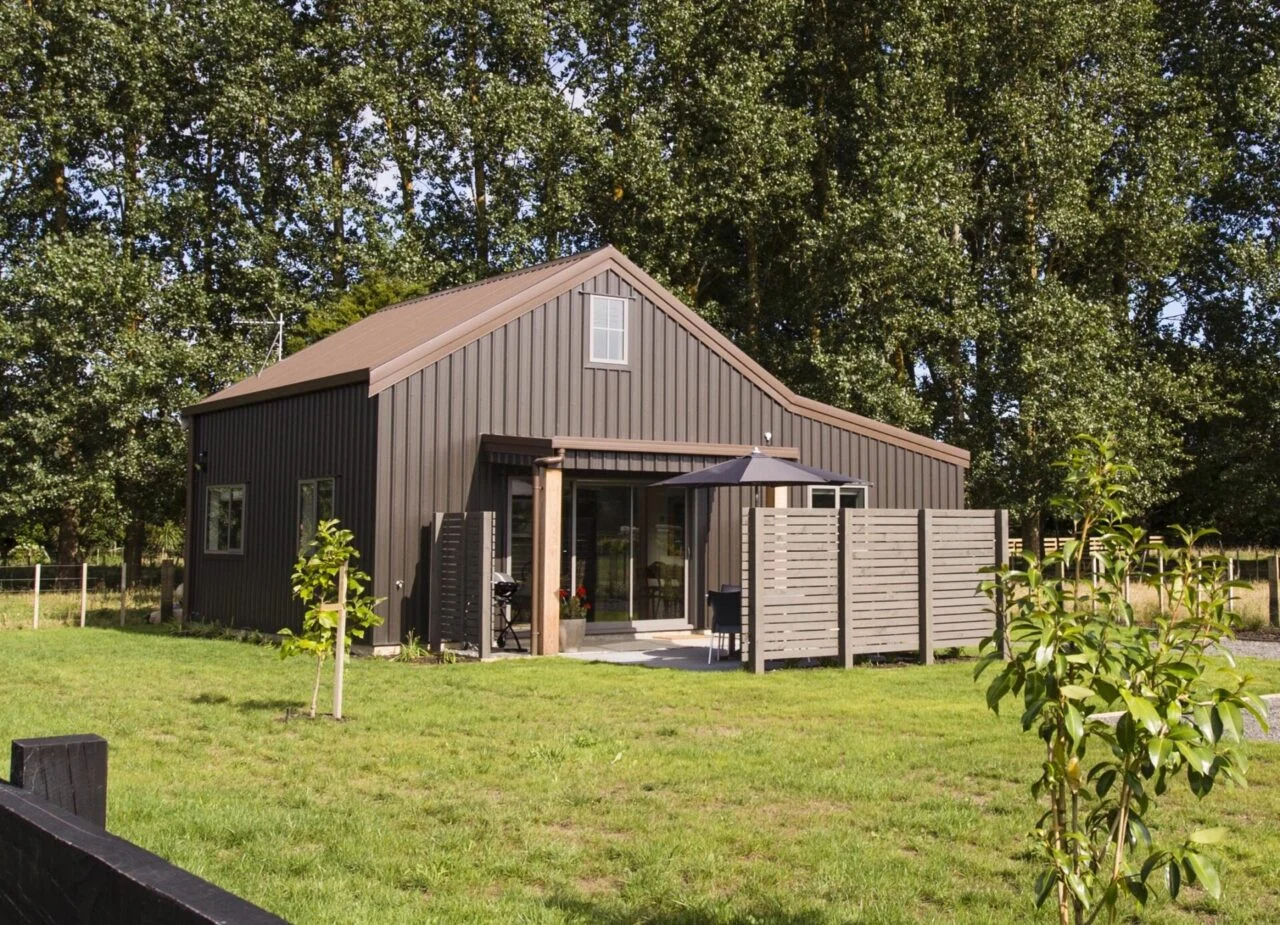 Brown steel-clad barn-style shed with a pitched roof, glazed doors, and a sheltered entrance in a green garden