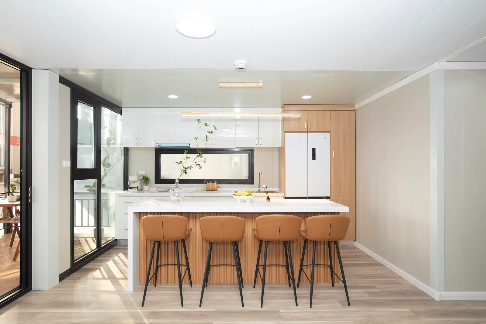 Sunlit Eco Portables kitchen with white cabinetry, a timber-fronted island, bar stools, and full-height windows